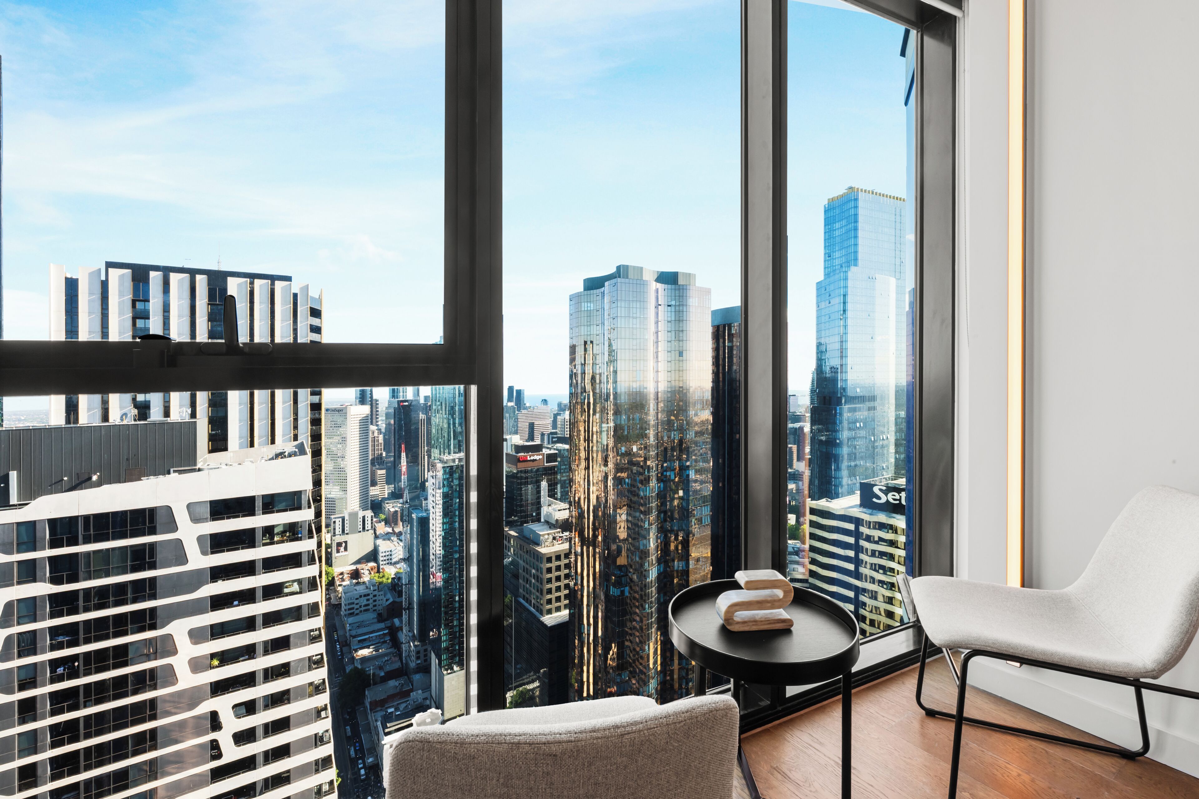 Ein modernes Zimmer mit großem Fenster, einem weißen Stuhl, einem kleinen runden Tisch mit Handtüchern und Blick auf eine Skyline mit hohen Gebäuden.