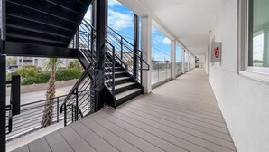 A covered outdoor corridor with a staircase, glass railings, and a fire extinguisher on the wall. - Brand New Oceanfront 2BR/2BA- Hot Tub, Heated Pool, EV Charger, 2 Parking spots (Carolina Beach)