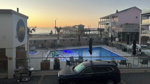 A pool area with a view of the ocean, a pier, and a building with a sign. - Brand New Oceanfront 2BR/2BA- Hot Tub, Heated Pool, EV Charger, 2 Parking spots (Carolina Beach)
