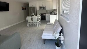 Una sala de estar moderna con sofá, una mesa de comedor con sillas, una zona de cocina con armarios y electrodomésticos, y una ventana con persianas.