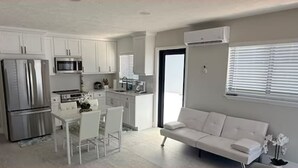 Cocina moderna con armarios blancos, refrigerador, estufa y microondas. Cuenta con un comedor con mesa y sillas, y un sofá blanco en la sala.