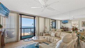 En rummelig opholdsstue med en stor sofa, et glasbord og havudsigt gennem den skydedør.