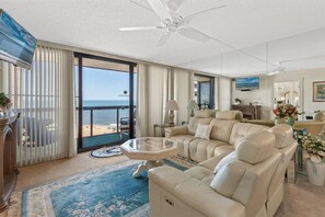 Un salón espacioso con un sofá grande, una mesa de centro de cristal y vista a la playa a través de puertas corredizas de vidrio.