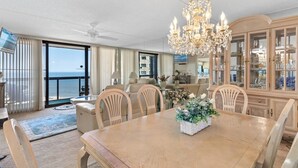 En rummelig stue med et stort spisebord, stole, en lysekrone og havudsigt.