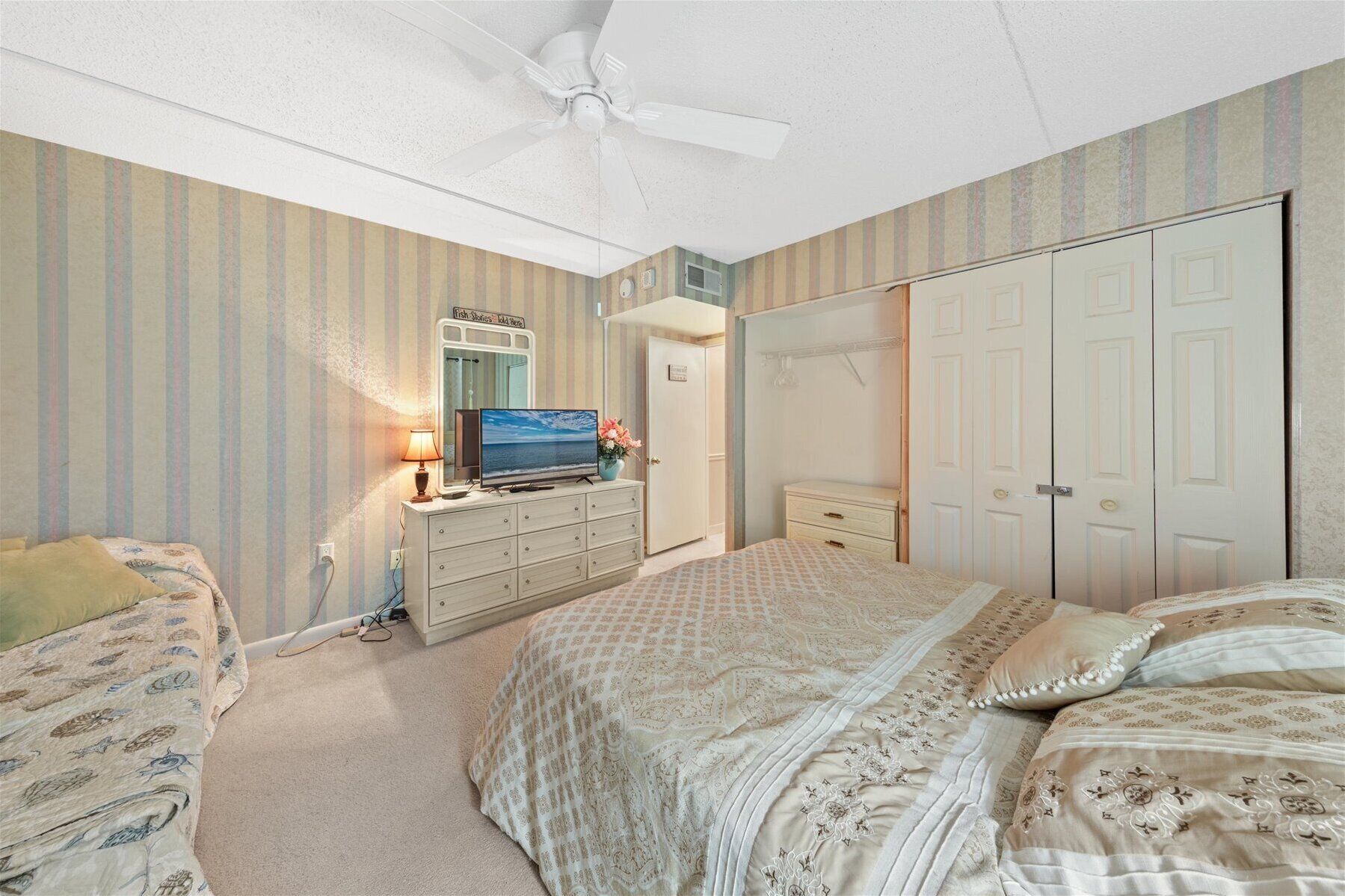 A bedroom with striped wallpaper, a ceiling fan, a television, a dresser, a bed with patterned bedding, and a wardrobe.