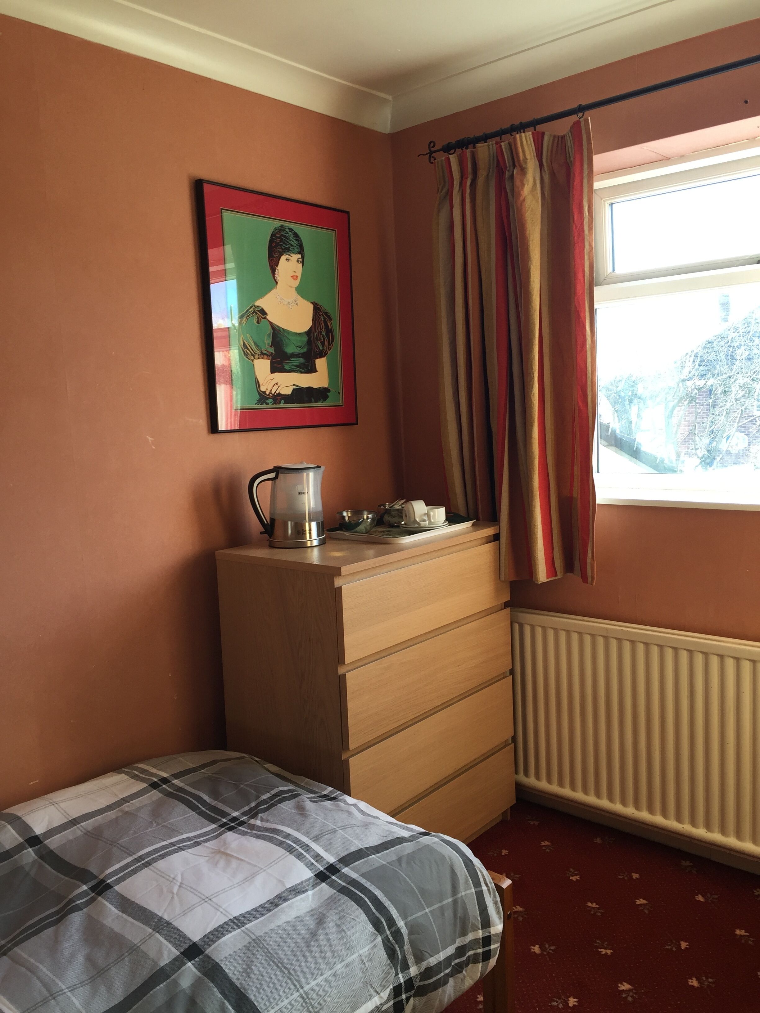 Un dormitorio con cama, cómoda, radiador, ventana con cortinas y un cuadro en la pared.