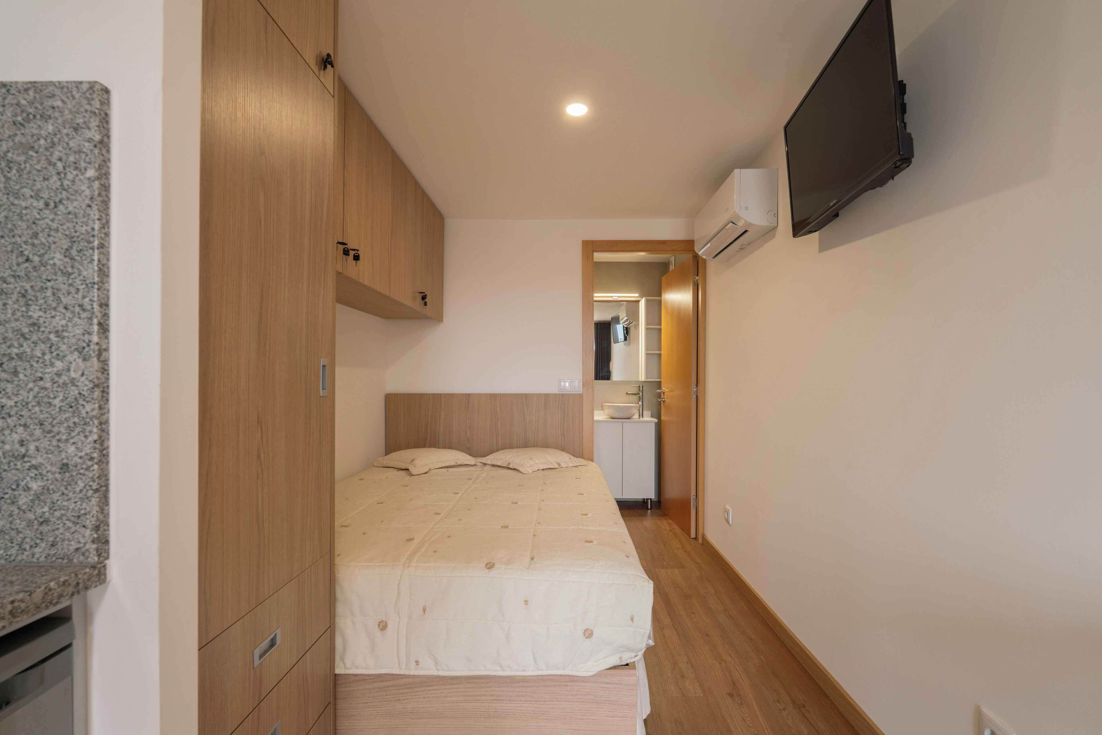 A bedroom with a bed, wooden wardrobe, built-in air conditioning unit, and a television mounted on the wall.