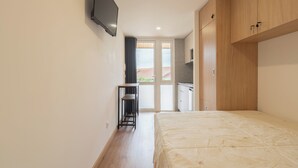 Un dormitorio con cama, televisión montada en la pared, una mesita y un área de kitchenette.