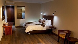 A hotel room with a wooden floor, a bed with white bedding, a desk, a chair, and a TV mounted on the wall. - Villa Sawah Hotel and Resort (Megamendung)