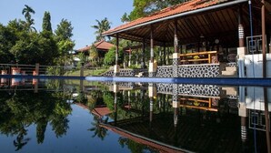 A building with a tiled roof, a covered outdoor area with a patterned floor, and a reflection in the water. - Villa Sawah Hotel and Resort (Megamendung)
