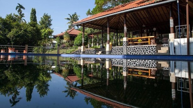 A building with a tiled roof, a covered outdoor area with a patterned floor, and a reflection in the water. - Villa Sawah Hotel and Resort (Megamendung)