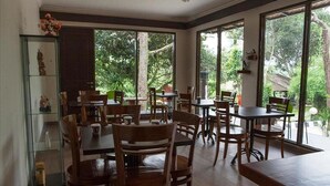 A spacious dining area with multiple tables and chairs, large windows offering a view of greenery, and a glass display case on the left. - Villa Sawah Hotel and Resort (Megamendung)