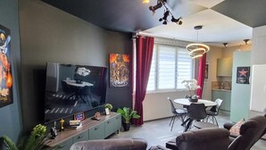 Ein modernes Wohnzimmer mit Flachbildfernseher, Esstisch mit Stühlen, einer kleinen Küchenzeile und einem großen Fenster mit Vorhängen.