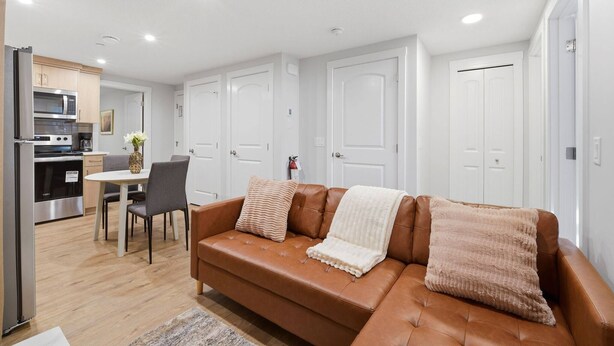 A modern living room with a brown leather sofa, a dining table with chairs, a kitchen area with a refrigerator and microwave, and white doors. - Luxury 2bedroom Suite With Park Views in Calgary (Calgary)