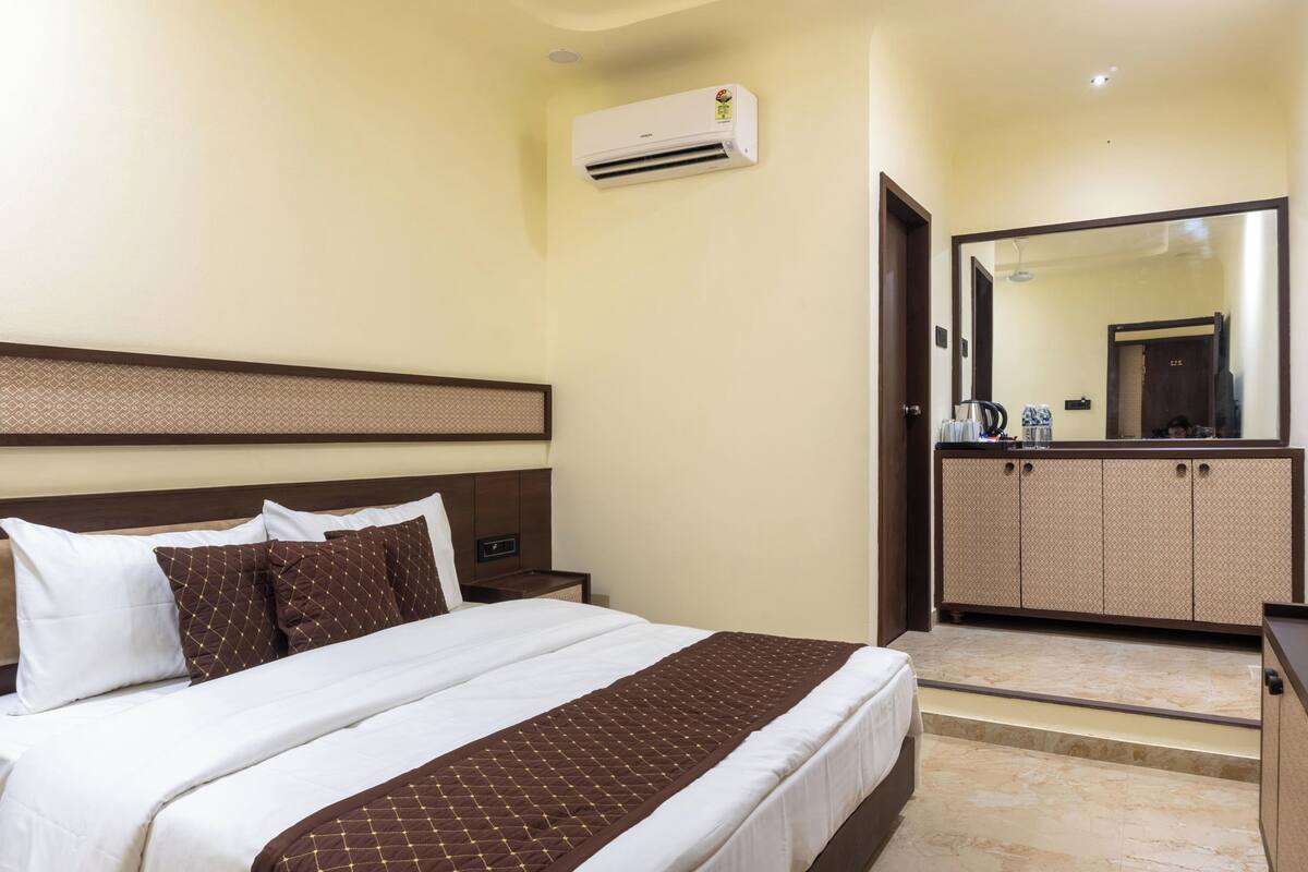 Habitación de hotel con cama, mesita de noche, equipo de aire acondicionado, espejo y zona de kitchenette.