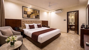 A hotel room with a large bed, a chair, a small table with a vase of flowers, a painting on the wall, and an air conditioning unit. - Hotel Liora (Rajkot)