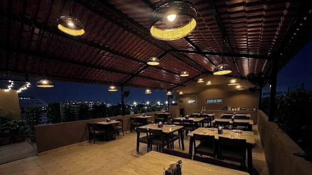 An outdoor dining area with multiple tables and chairs, a high ceiling with hanging lights, and a city view in the background. - Hotel Liora (Rajkot)