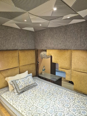 Un dormitorio con una colcha estampada, un cabecero con ventilador incorporado, una mesita de noche con una botella y un espejo.