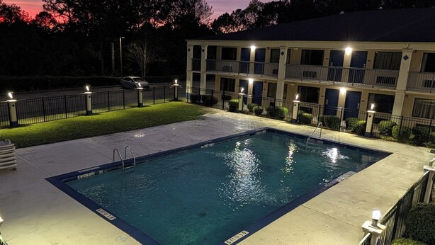 Una piscina rectangular con un paseo alrededor, un edificio con varios balcones y una valla.