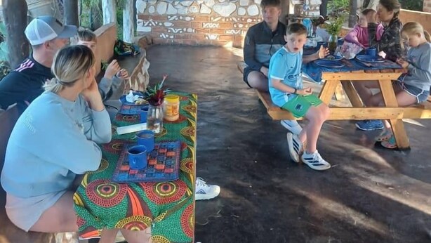 Un área de comedor al aire libre con mesas y bancos de madera, rodeada de muros de piedra y un techo de paja.