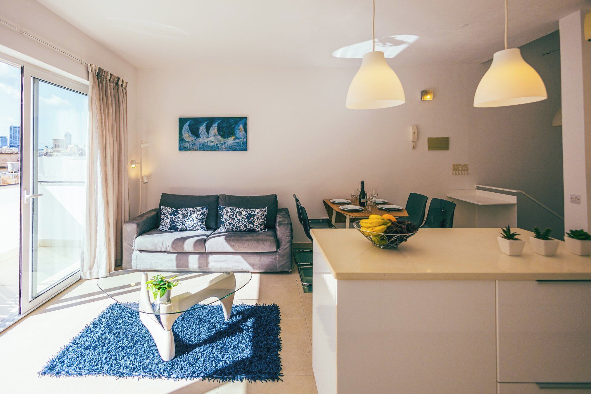 A modern living room with a sofa, a dining table with chairs, a glass coffee table, and a kitchen area with a counter and plants.