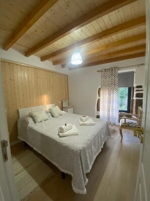 Ein Schlafzimmer mit Holzbalkendecke, einem Bett mit weißer Bettwäsche, einem Nachttisch, einem Sessel und einem Fenster mit Vorhängen.