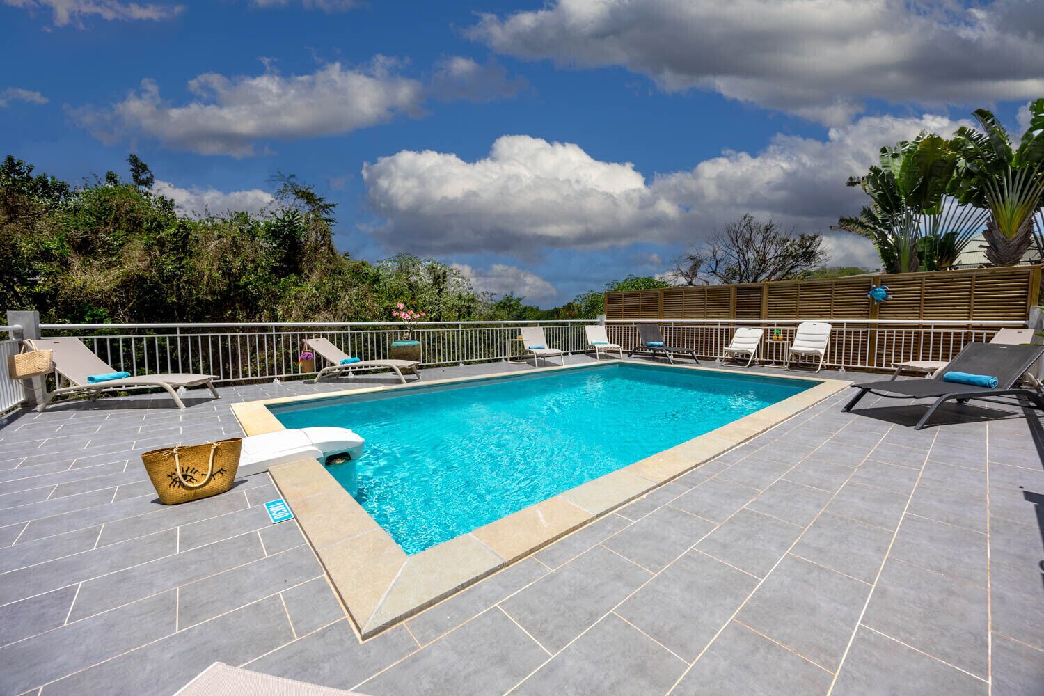A rectangular pool with a tiled deck, surrounded by lounge chairs and a wooden fence.