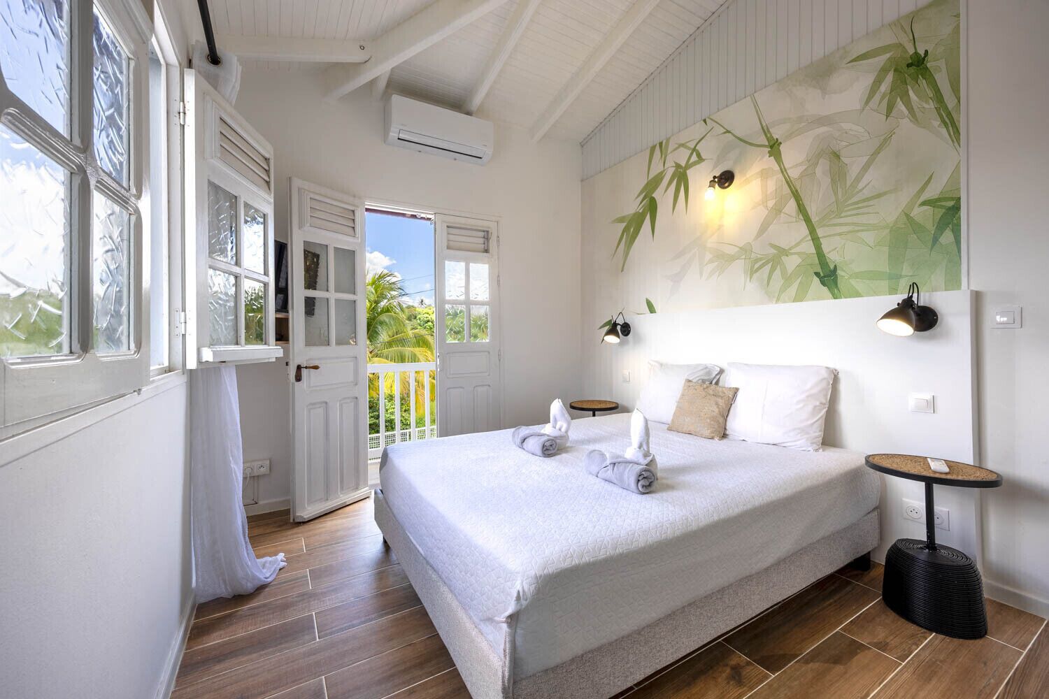 A bedroom with a bed, a nightstand, a wall-mounted lamp, and a view of the outdoors.