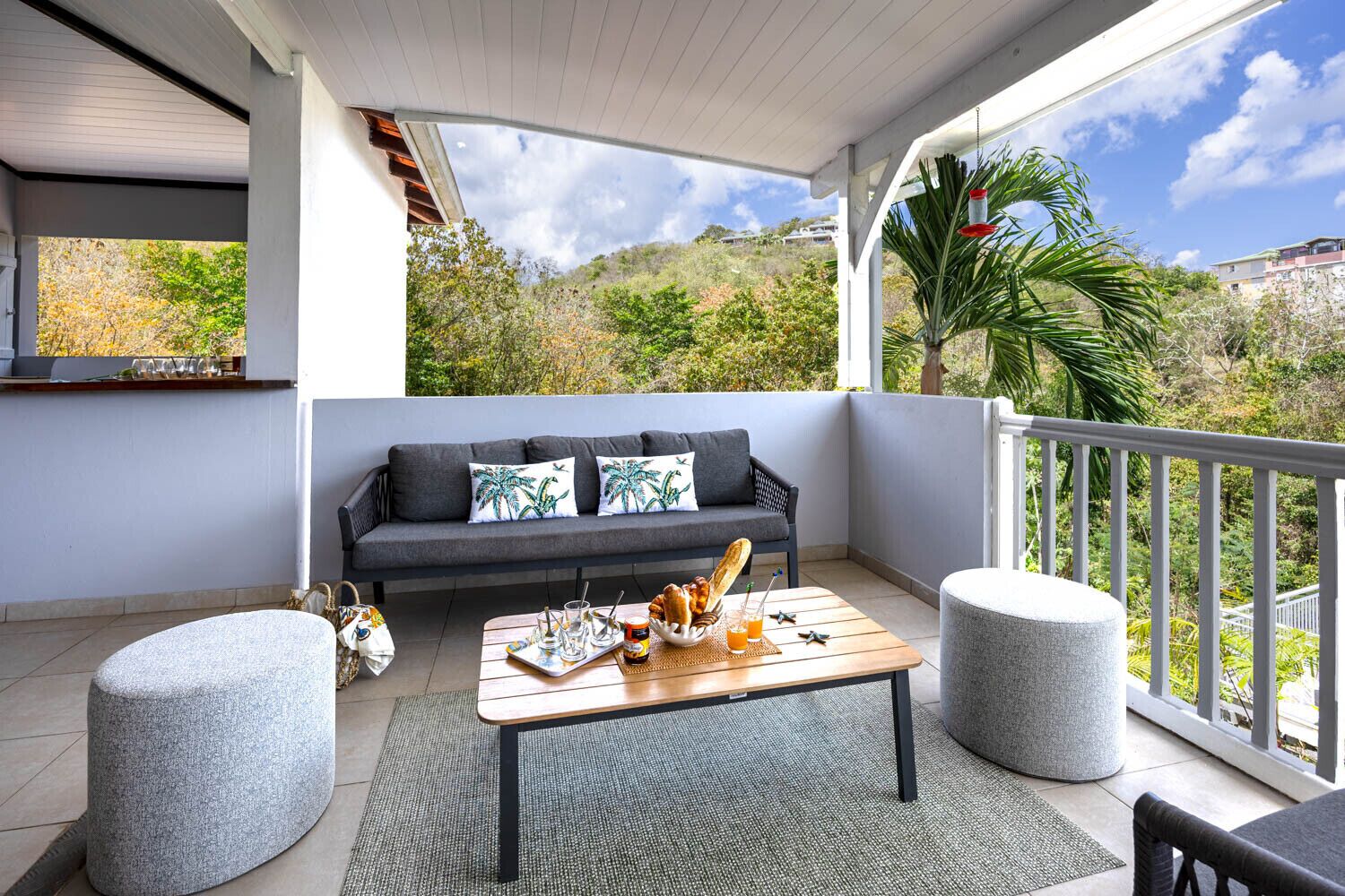 A covered balcony with a sofa, two ottomans, a wooden table with breakfast items, and a view of greenery and buildings.