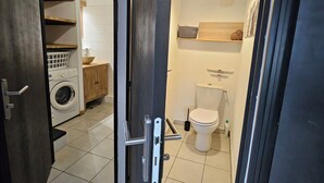 Eine geöffnete Tür zu einem Waschraum mit Waschmaschine und Regalen, einer Toilette mit Bidet sowie einer Pendelleuchte.