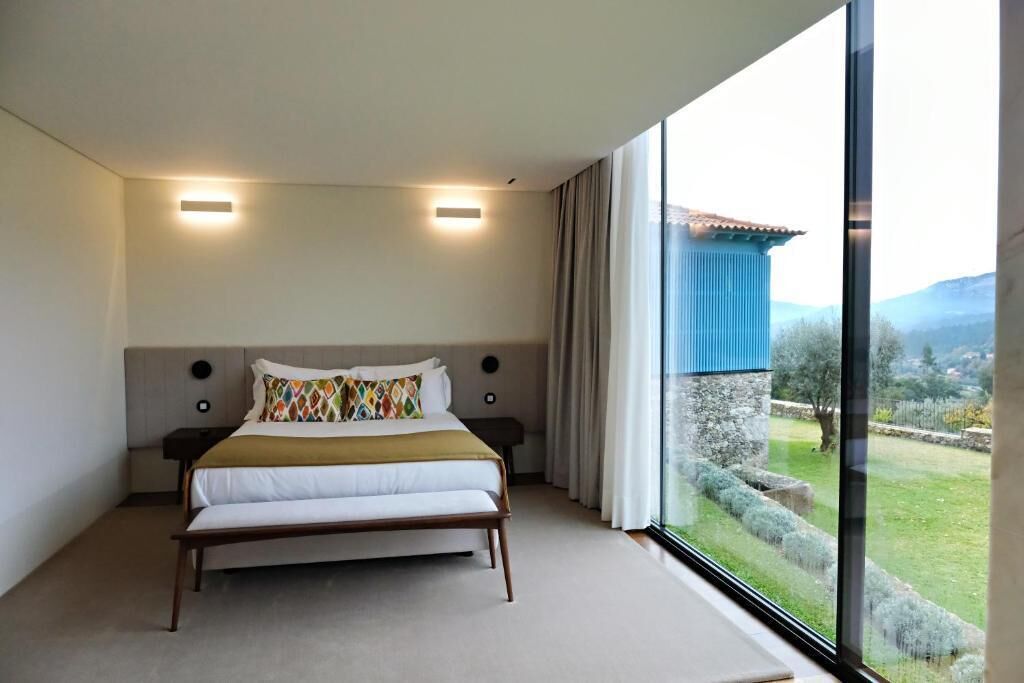 A bedroom with a large bed, two bedside lamps, a view of a garden, and a window with curtains.