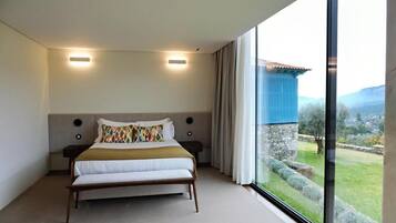 A bedroom with a large bed, two bedside lamps, a view of a garden, and a window with curtains.