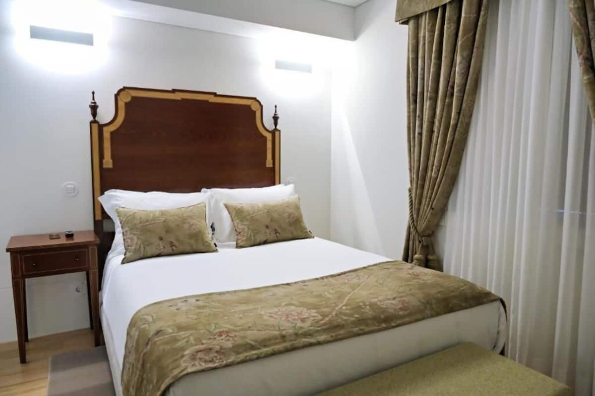 Un dormitorio con cabecera de madera, cama con ropa de cama blanca, una mesita de noche y una ventana con cortinas.
