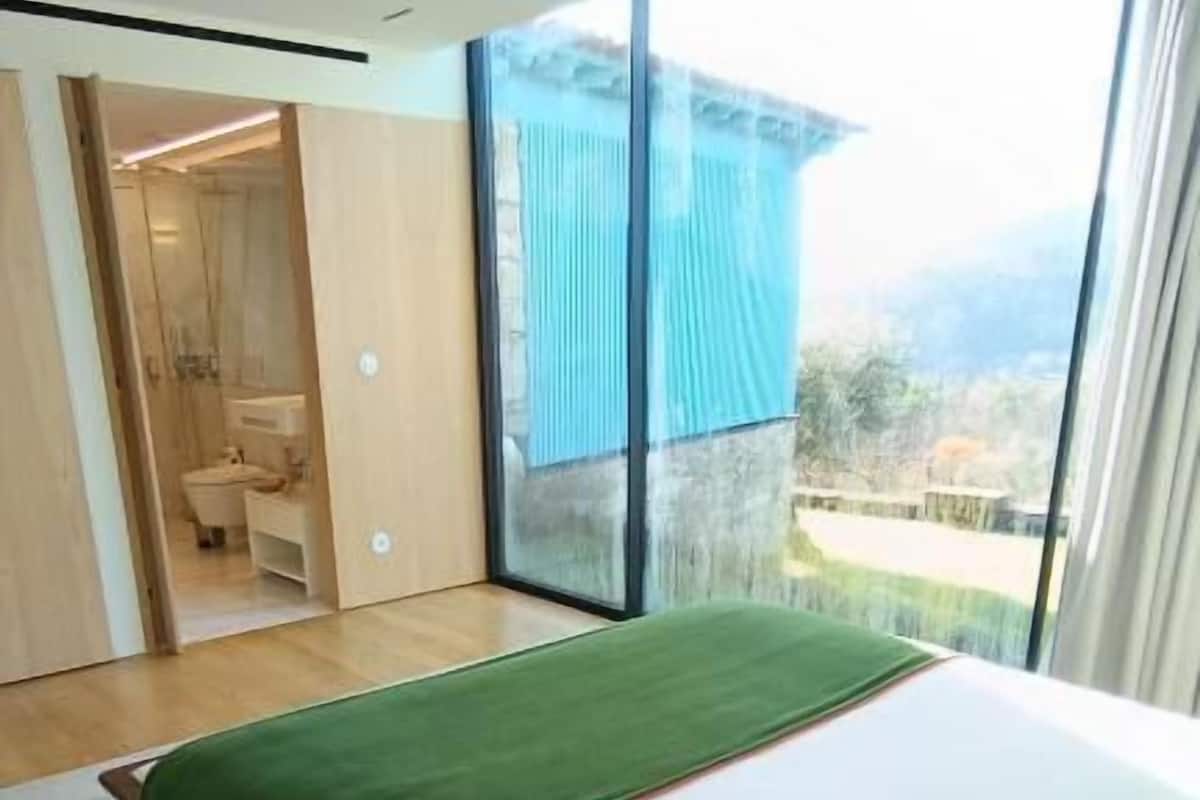 Un dormitorio con cama, una manta verde, una ventana y un baño visible a través de una puerta abierta.