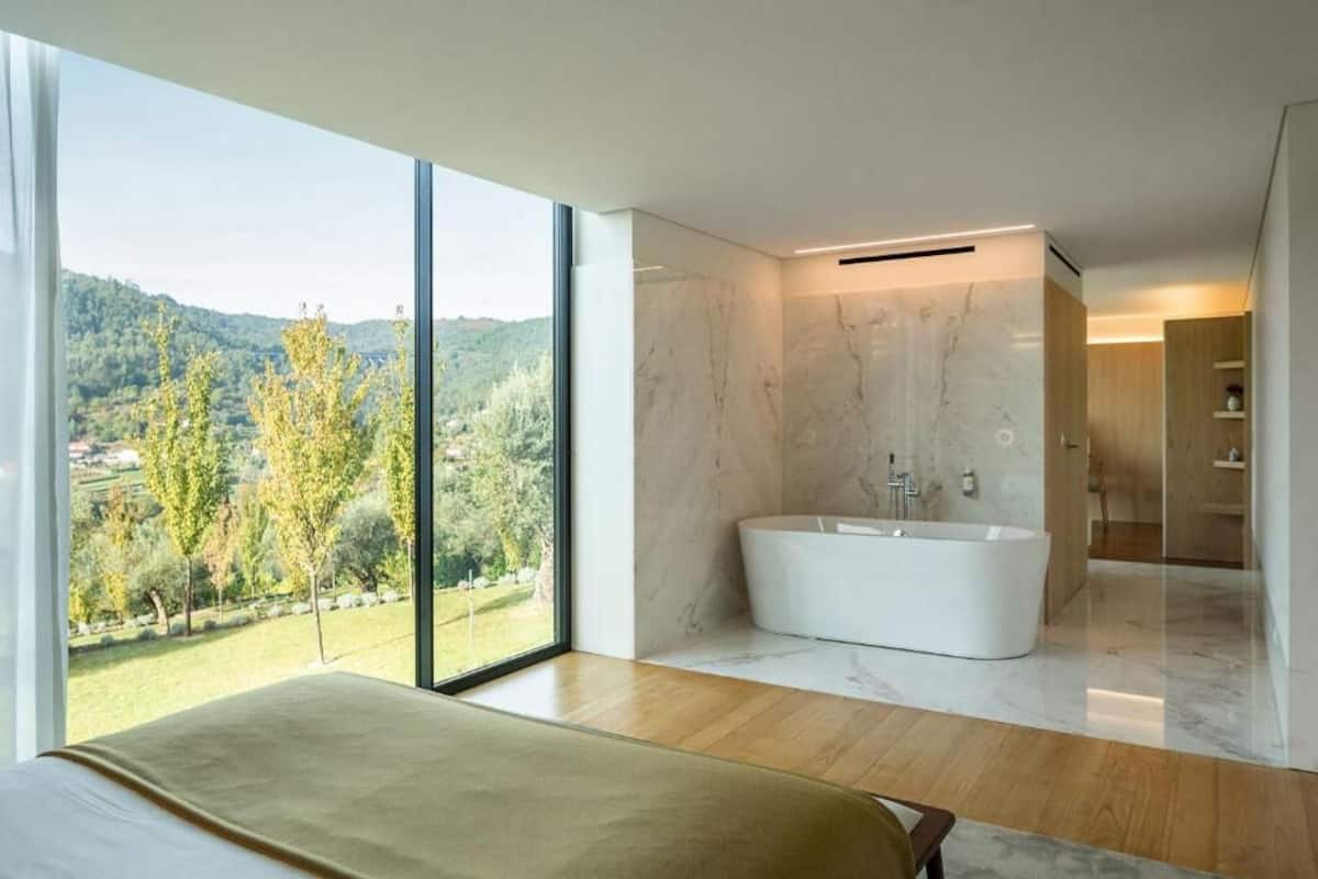 Un baño moderno con una bañera independiente, un ventanal con vistas a un paisaje verde y suelo de madera.