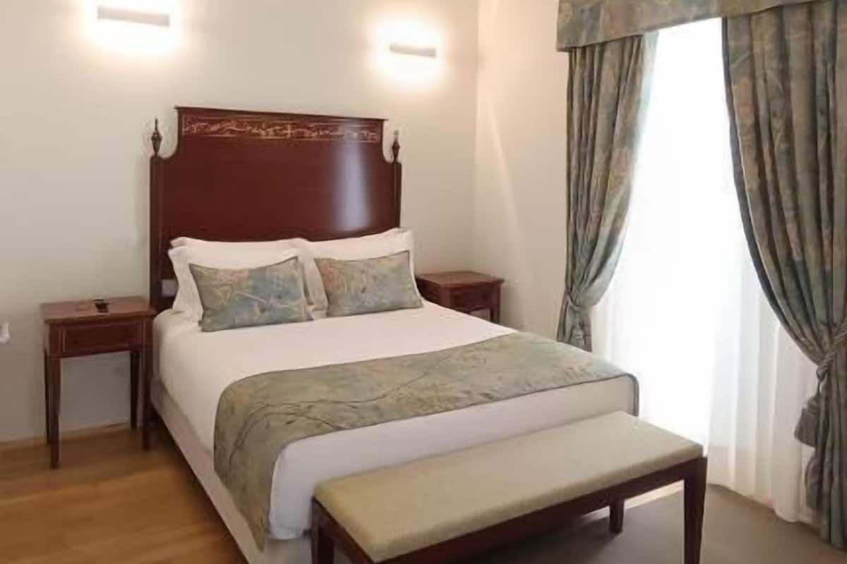 Un dormitorio con cama de madera, mesitas de noche, un banco, techo inclinado y una ventana con cortinas.