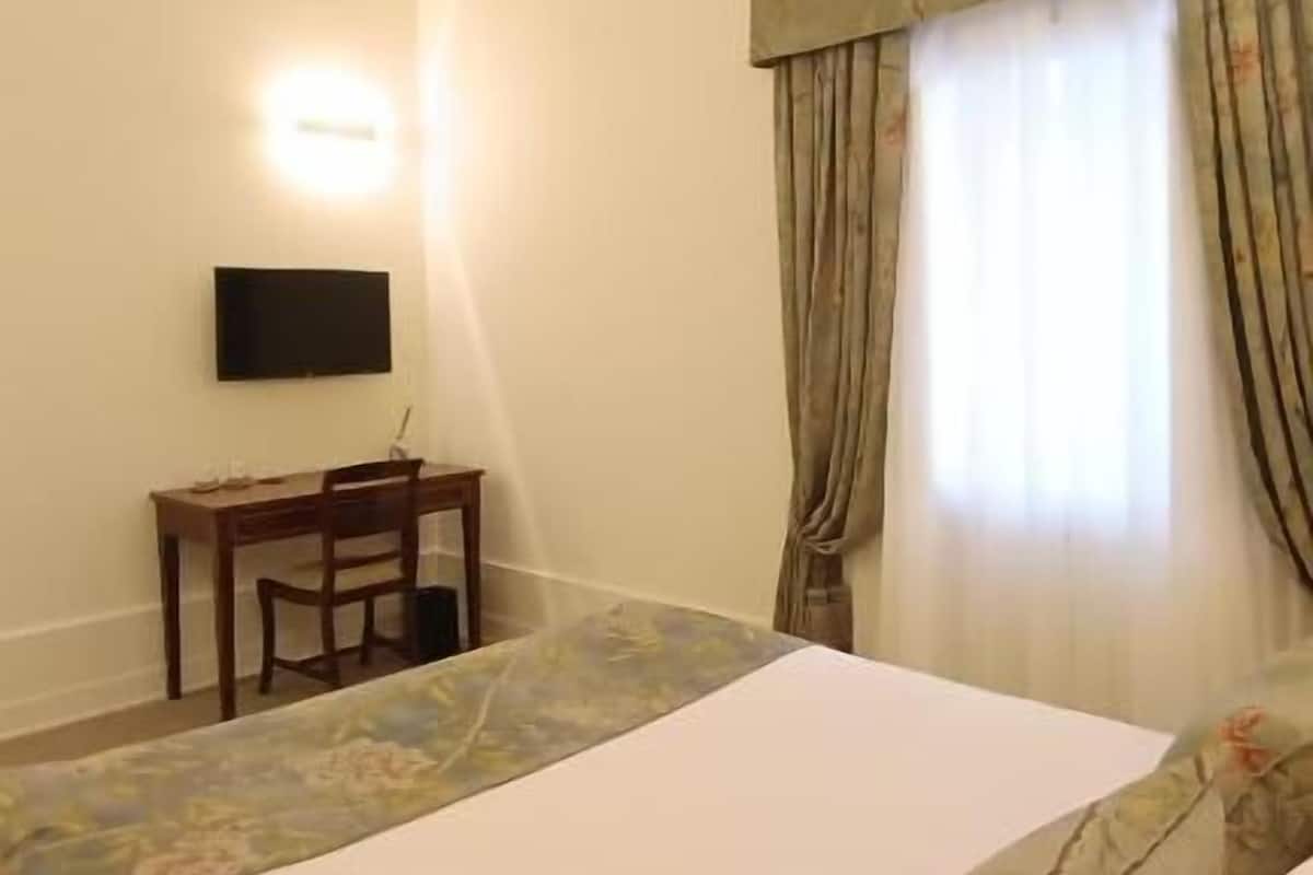 Habitación de hotel con cama, escritorio, silla, televisor y ventana con cortinas.