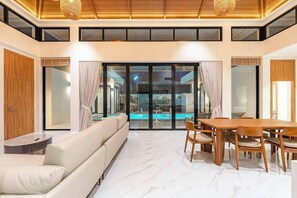 Ein modernes Interieur mit einer Couch, einem Esstisch, Stühlen, großen Fenstern und Blick auf einen Swimmingpool.