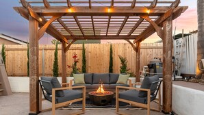 A wooden pergola with string lights, a fire pit, and a sofa with chairs in a backyard setting. - Resort-Style Pool Retreat | Home Gym + Hot Tub (Goleta)