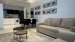 Ein modernes Wohnzimmer mit einer Couch, einem Couchtisch und einem Essbereich mit Stühlen. Im Hintergrund befindet sich eine Küche mit Waschmaschine und Kühlschrank.