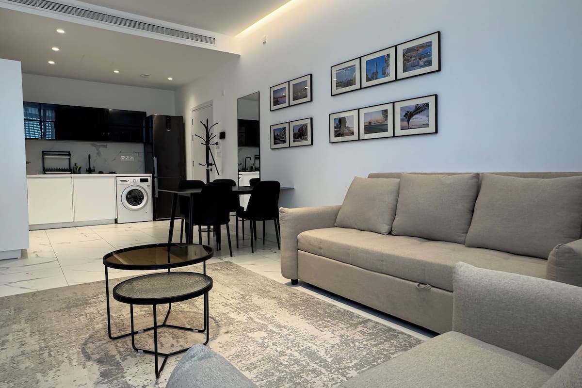 Una sala moderna con un sofá, una mesa de centro y un comedor con sillas. Se observa una cocina al fondo con lavadora y refrigerador.