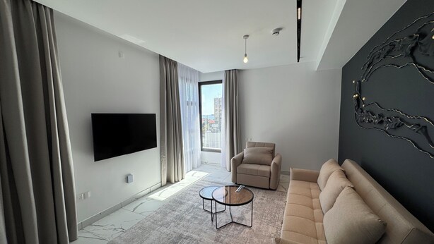 Ein modernes Wohnzimmer mit Flachbildfernseher, einer Couch, einem Couchtisch und einem großen Fenster mit Vorhängen.
