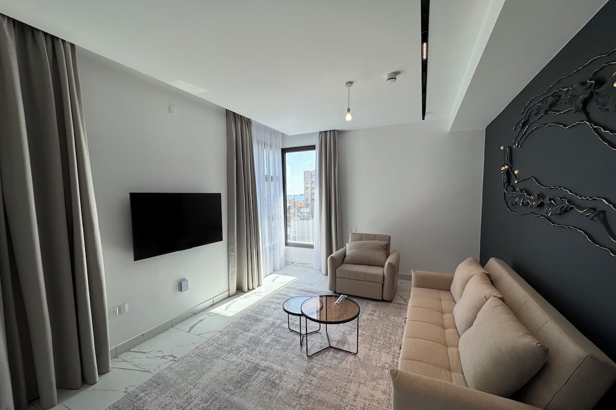 Una sala de estar moderna con un televisor de pantalla plana, un sofá, una mesa de centro y un amplio ventanal con cortinas.
