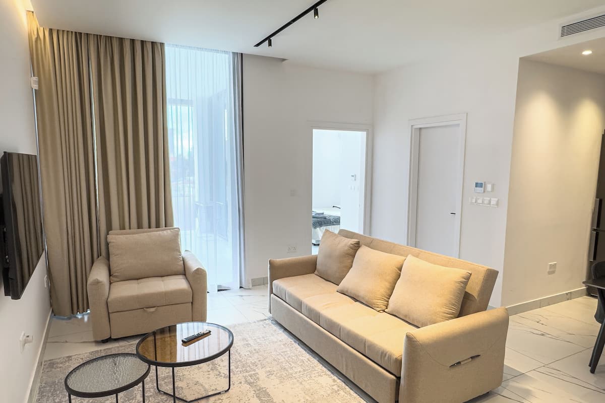 Una sala de estar moderna con un sofá, un sillón y una mesa de centro, una ventana grande con cortinas y una puerta que da a otra habitación.