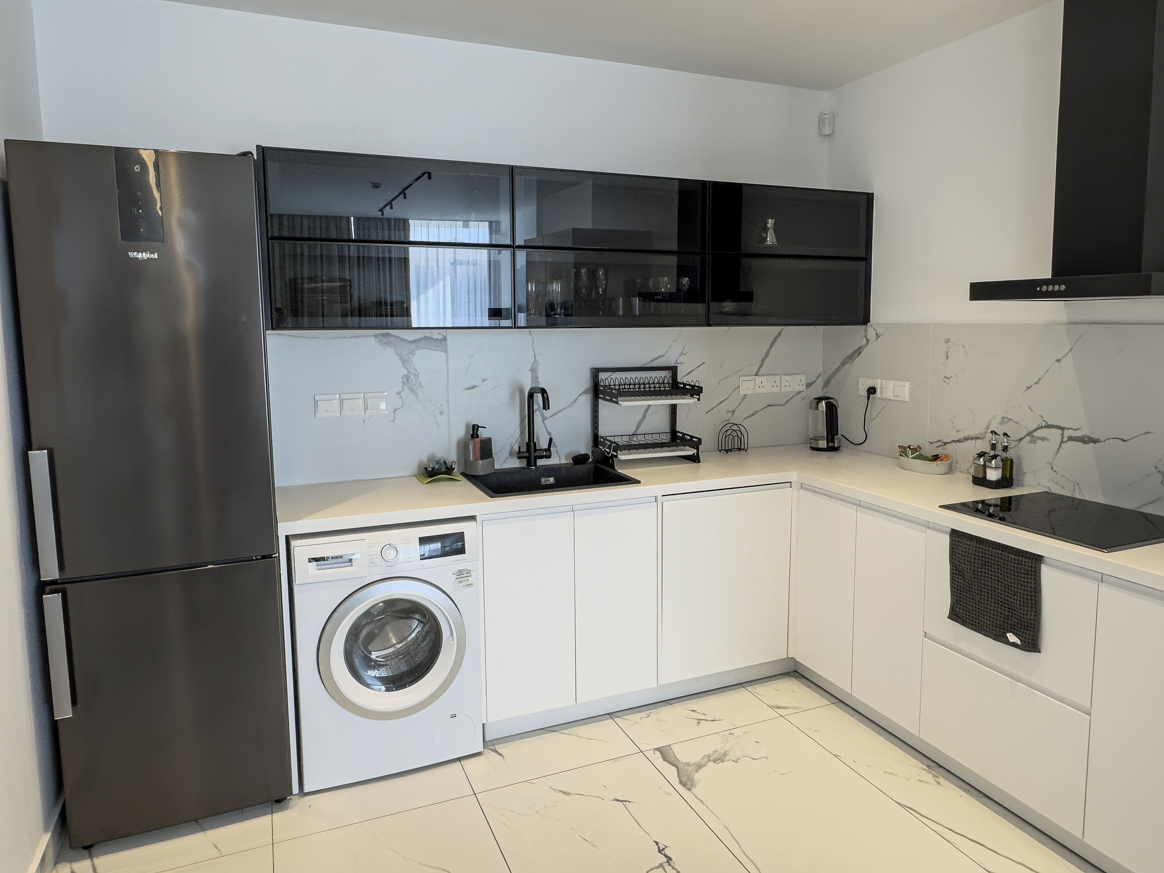 A modern kitchen with a stainless steel refrigerator, a built-in washing machine, a black countertop, and white cabinets.