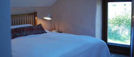 A bedroom with a bed, a bedside lamp, a window with a view of greenery, and a sloped ceiling.