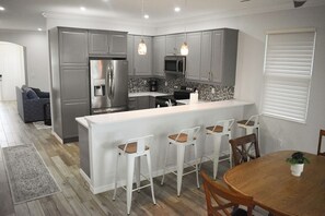 Eine moderne Küche mit grauen Schränken, einem Edelstahlkühlschrank, einer Mikrowelle und einer Kaffeemaschine. Es gibt eine weiße Kücheninsel mit Barhocker und einen Holzessttisch mit Stühlen.