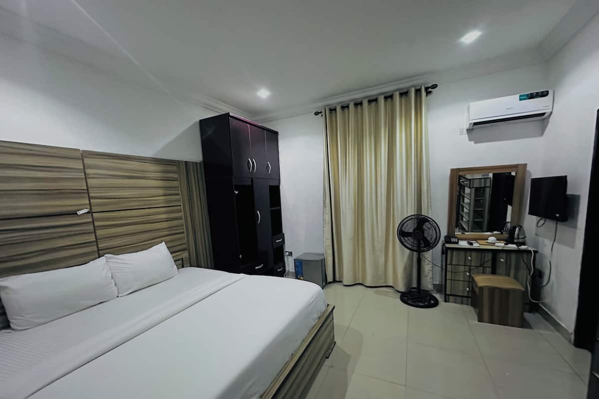 A hotel room with a bed, wardrobe, air conditioning unit, fan, and a desk with a mirror and a speaker.