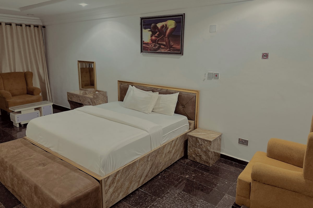 A hotel room with a large bed, a chair, a vanity with a mirror, and a picture on the wall.