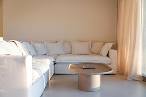 A modern living room with a sectional sofa, a round coffee table, and a large window with curtains. - Beach House Houlakia Mykonos (Mykonos)